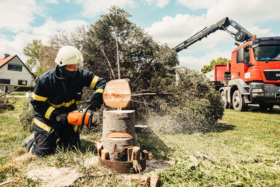 A firefighter wearing protective gear, including a helmet and gloves, is kneeling on the grass while operating a chainsaw to cut a large tree trunk into sections. In the background, a crane truck with