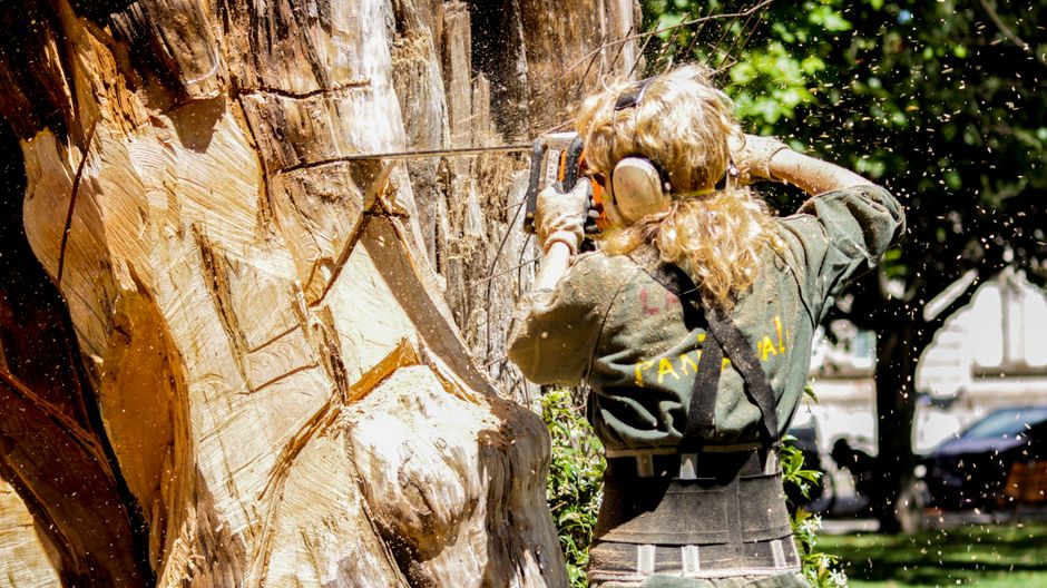 A firefighter wearing protective gear, including a helmet and gloves, is kneeling on the grass while operating a chainsaw to cut a large tree trunk into sections. In the background, a crane truck with